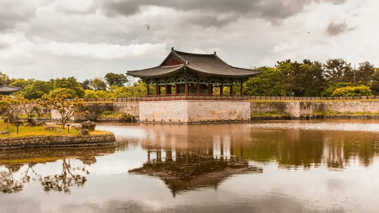 A traditional Korean pavilion on a stone foundation sits over a calm lake, its reflection visible in the water under a cloudy sky.