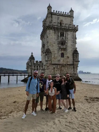 Un gruppo WeRoad di sette persone in posa su una spiaggia di sabbia davanti a una grande torre di pietra in riva al mare.
