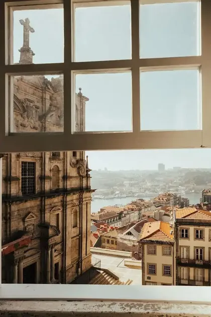 Veduta di una storica città europea con tetti rossi e una chiesa in pietra, osservata dall'interno attraverso una finestra a più riquadri.