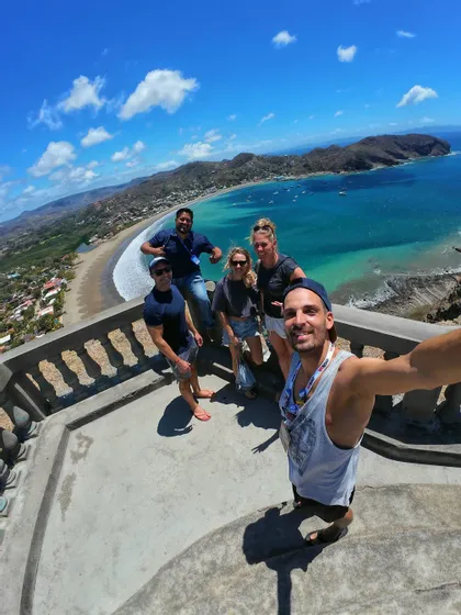 Eine WeRoad-Gruppe macht ein Weitwinkel-Selfie auf einem Aussichtspunkt mit Blick auf eine malerische Bucht mit einem Sandstrand und Hügeln.