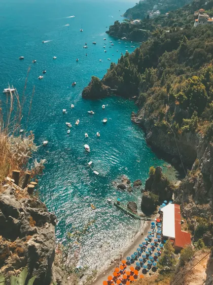 Veduta dall'alto di una costa rocciosa con una caletta dalle acque turchesi, numerose imbarcazioni e una piccola spiaggia con file di ombrelloni colorati.