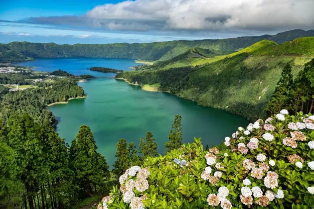 Una vista panoramica di due laghi di caldera collegati, uno verde e uno blu, circondati da lussureggianti colline boscose con ortensie in primo piano.