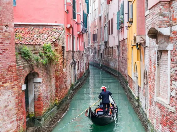 Un gondolier vu de dos rame une gondole le long d'un étroit canal entre des bâtiments historiques en briques colorées.