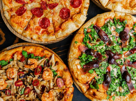 Vue plongeante de trois pizzas différentes aux garnitures variées, telles que pepperoni, crevettes et roquette, présentées sur une surface sombre.