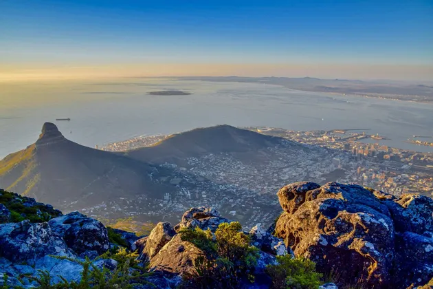 Une ville côtière et un pic montagneux vus d'un point de vue rocheux élevé sous un ciel clair au lever du soleil.