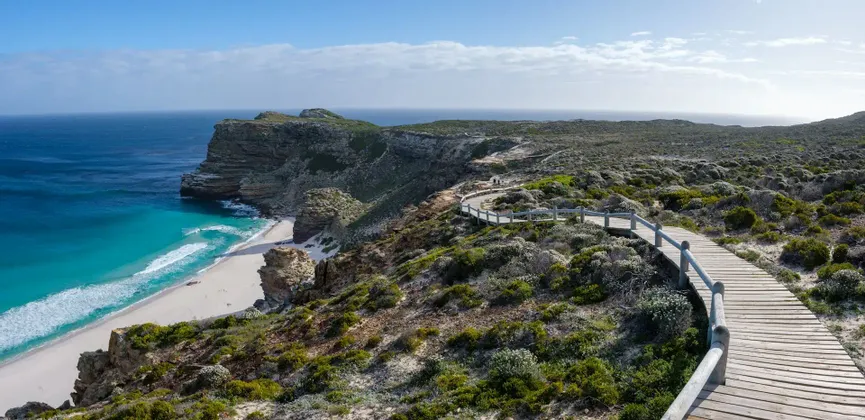 Une passerelle en bois serpente le long d'une falaise escarpée et verdoyante, surplombant une plage de sable blanc et un océan turquoise sous un ciel bleu.