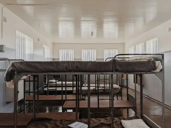 Intérieur d'un dortoir austère avec des rangées de lits superposés en métal, des couvertures en laine et des fenêtres à barreaux.