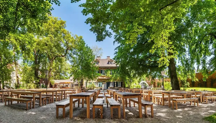 File di tavoli e panche di legno vuoti si trovano su un patio ghiaioso in una birreria all'aperto, ombreggiata da grandi alberi frondosi.