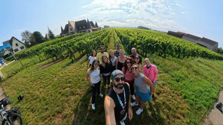 Un gruppo WeRoad si scatta un selfie in un vigneto soleggiato con filari di viti e un castello sullo sfondo.