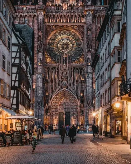Una veduta di una strada acciottolata al crepuscolo che conduce alla facciata illuminata di una grande e ornata cattedrale gotica con un imponente rosone.