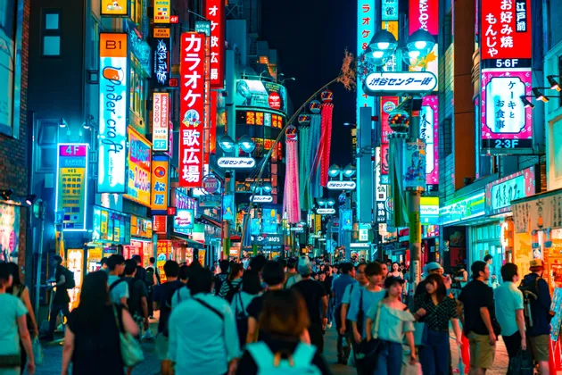Eine große Menschenmenge geht nachts eine belebte Stadtstraße entlang, hell erleuchtet von hoch aufragenden Neonschildern mit japanischen Schriftzeichen.
