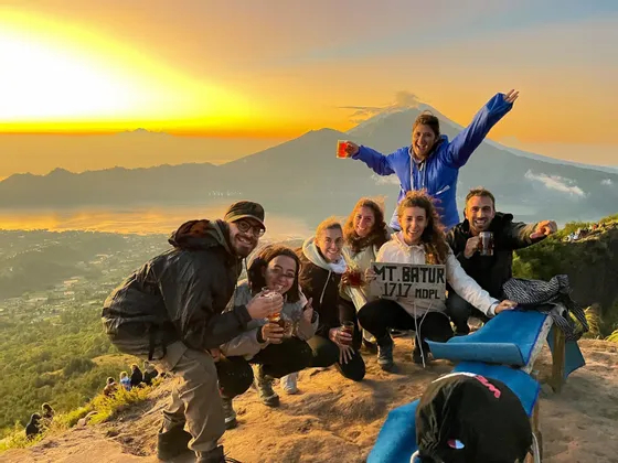 Eine WeRoad-Gruppe lächelt für ein Foto auf einem Berggipfel, hält Getränke und ein Schild für den Mt. Batur bei Sonnenaufgang.