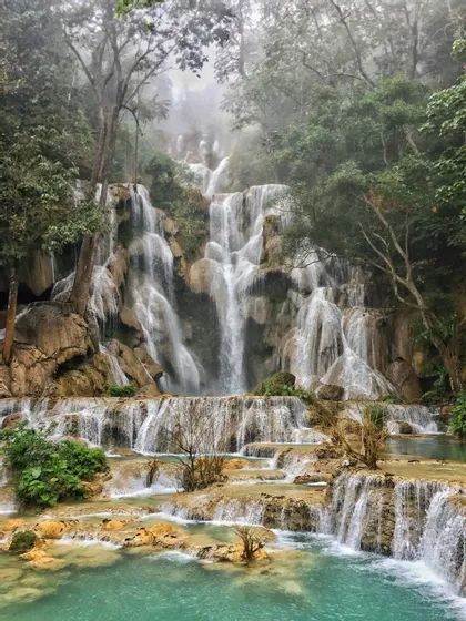 Una cascata a gradoni scende da ripiani rocciosi in pozze turchesi, circondata da una foresta lussureggiante e nebbiosa.