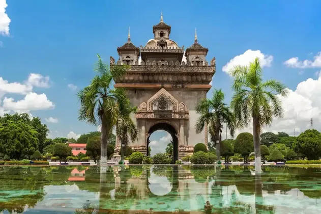 Un arco in pietra finemente decorato, riflesso in una piscina, circondato da palme e giardini sotto un cielo azzurro.