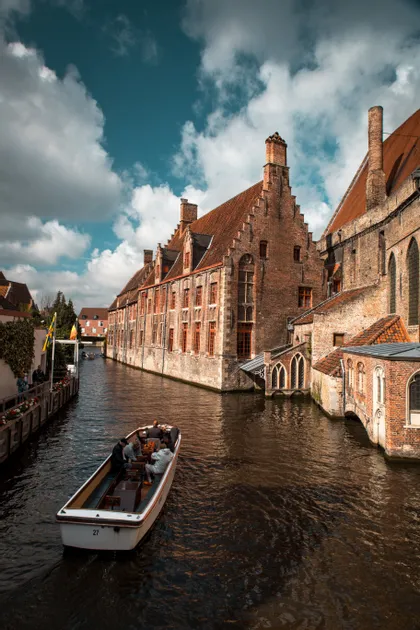 Ein Ausflugsboot mit Passagieren fährt auf einem Kanal entlang, vorbei an historischen Backsteingebäuden mit Giebeldächern, unter bewölktem Himmel.