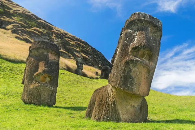 Diverse grandi statue Moai di pietra si ergono su una collina erbosa sotto un cielo azzurro brillante.