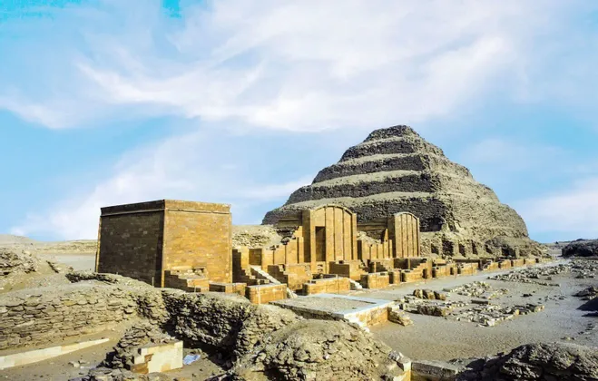 Une grande pyramide à degrés se dresse parmi des ruines en pierre anciennes dans un paysage désertique sous un ciel bleu partiellement nuageux.