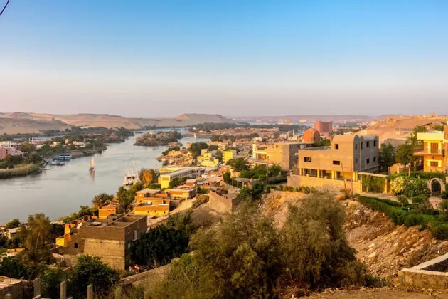 Vue en plongée d'une ville aux bâtiments couleur sable le long d'une large rivière avec des voiliers, sur fond de collines désertiques.