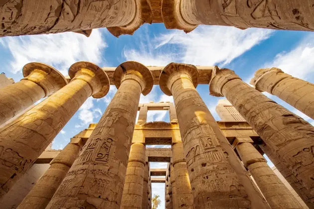 Une vue en contre-plongée de colonnes antiques massives avec des gravures hiéroglyphiques s'élevant vers un ciel bleu parsemé de nuages blancs.