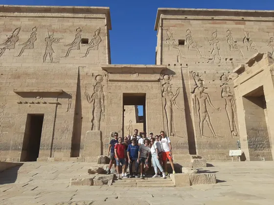 Un groupe WeRoad pose pour une photo sur les marches d'un grand temple égyptien antique orné de hiéroglyphes, sous un ciel bleu clair.