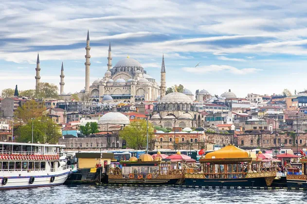 Eine große Moschee mit Kuppeln und Minaretten überblickt eine städtische Uferpromenade, an der mehrere Boote festgemacht sind.