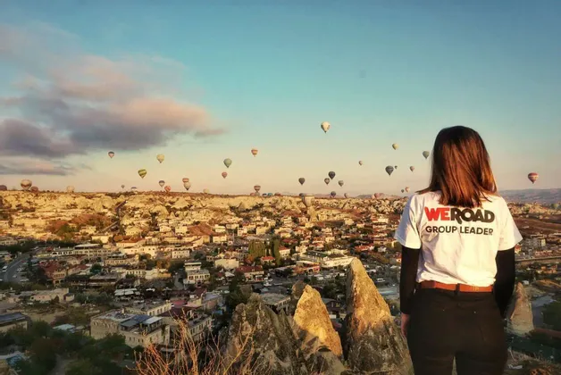 Ein WeRoad-Gruppenleiter beobachtet von hinten, wie Heißluftballons bei Sonnenaufgang über einer Stadt in einer felsigen Landschaft fliegen.