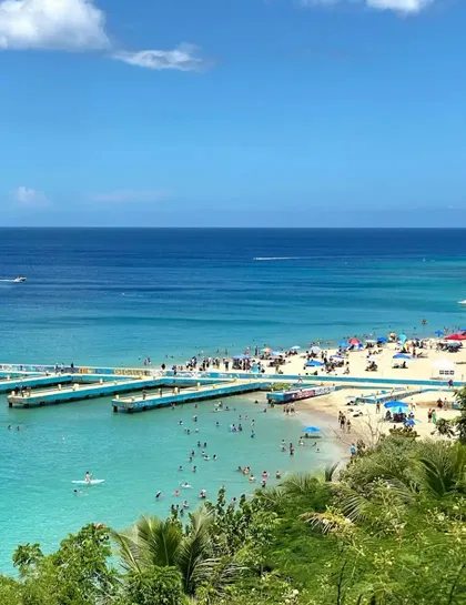Una vista aérea de una playa concurrida donde la gente nada en aguas turquesas y se relaja en la arena junto a largos muelles azules.