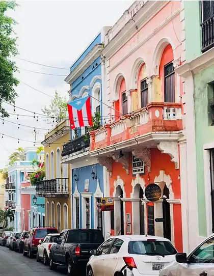 Una bandera puertorriqueña cuelga de un balcón en una calle llena de edificios coloniales coloridos y coches aparcados.