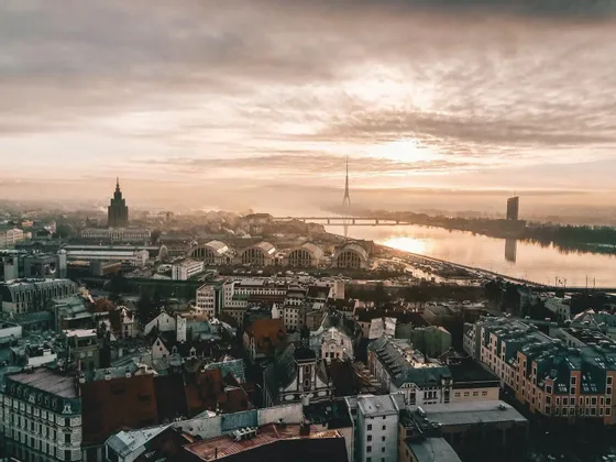 Una vista aerea dello skyline di una città accanto a un fiume largo durante un'alba nebbiosa, con vari edifici e un'alta torre in lontananza.