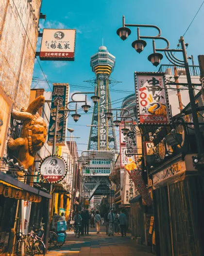 A view down a narrow city street crowded with Japanese signs, with a tall observation tower in the distance under a blue sky.