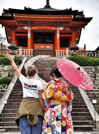 A WeRoad group leader makes a peace sign next to a woman in a kimono at the base of stairs leading to a Japanese temple.