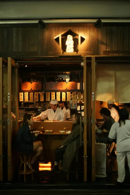 Two women on a WeRoad group trip dining at a traditional Japanese restaurant at night, viewed from the outside.
