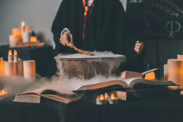 Una persona in tunica nera punta una bacchetta magica su un calderone fumante su un tavolo con libri aperti e candele accese.