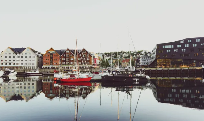 Boote liegen in einem ruhigen Hafen vor Anker, während sich traditionelle und moderne Gebäude entlang der Uferpromenade im Wasser spiegeln.
