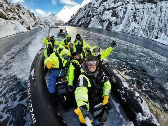 Eine Person macht ein Selfie während einer WeRoad Gruppenreise auf einem Festrumpfschlauchboot, das durch einen Fjord mit schneebedeckten Bergen fährt.