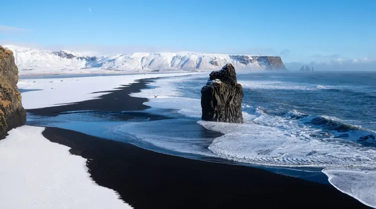 Olas rompen en una playa de arena negra parcialmente cubierta de nieve, con un gran farallón marino en la costa y montañas nevadas en la distancia.