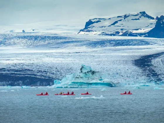 Un viaje en grupo de WeRoad rema en kayaks rojos en un lago glacial, con un glaciar enorme y montañas nevadas de fondo.