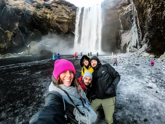 Un viaje en grupo de WeRoad de cuatro personas con ropa de invierno se toma una selfie junto a una cascada en un terreno nevado de arena negra.
