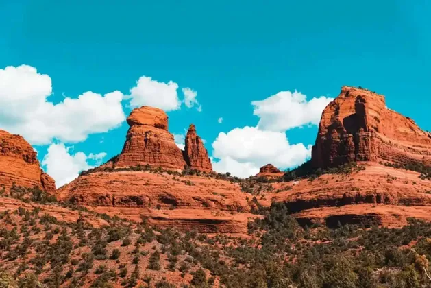 Geschichtete rote Felsformationen erheben sich aus einer Landschaft grüner Sträucher unter einem strahlend blauen Himmel mit bauschigen weißen Wolken.