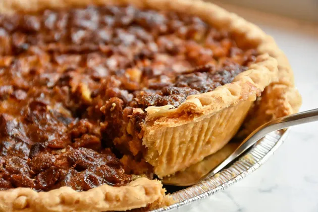 A close-up of a fork lifting a slice from a pecan pie with a flaky crust in a foil pan.