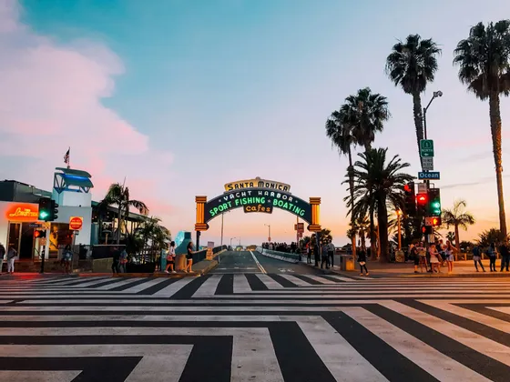 A street view of the illuminated Santa Monica Pier entrance sign, with a crosswalk in the foreground and palm trees at sunset.
