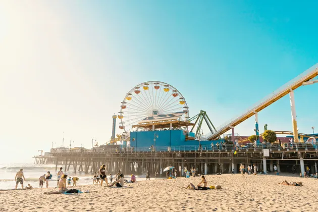 People relax on a sandy beach in front of a pier holding an amusement park with a Ferris wheel and roller coaster.