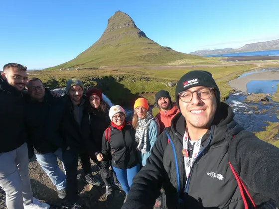 Eine WeRoad-Gruppenreise macht ein Selfie lächelnd vor einem großen grünen Berg und einem Fluss an einem sonnigen Tag.