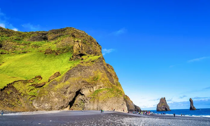 Eine WeRoad Gruppenreise an einem schwarzen Sandstrand neben einer moosbewachsenen Klippe, mit im Meer sichtbaren Felsnadeln unter klarem blauem Himmel.