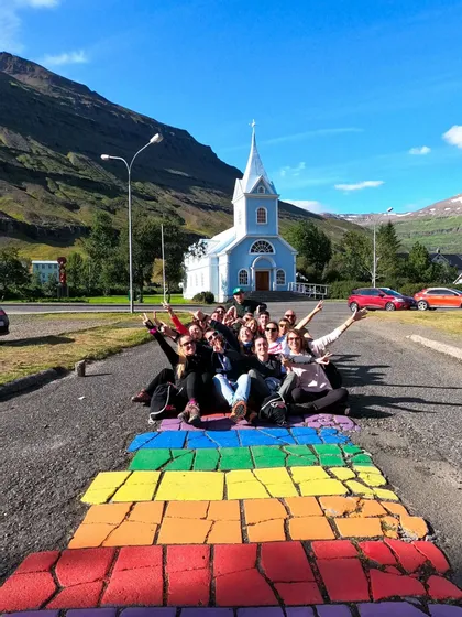 Eine WeRoad-Reisegruppe posiert auf einem regenbogenfarbenen Weg vor einer blauen Kirche und Bergen für ein Foto.