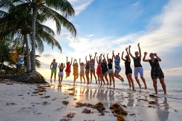 Un viaggio di gruppo WeRoad che salta insieme su una spiaggia tropicale al tramonto, con palme lungo la riva.