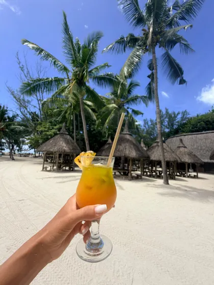 Une main tenant un cocktail tropical sur une plage de sable blanc avec des palmiers et des cabanes au toit de chaume en arrière-plan.