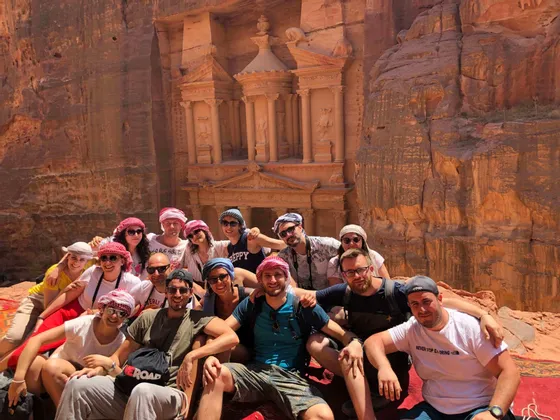 Eine WeRoad-Gruppe lächelt für ein Foto vor einem großen, in eine sonnenbeschienene, rötliche Felswand gehauenen Tempel.