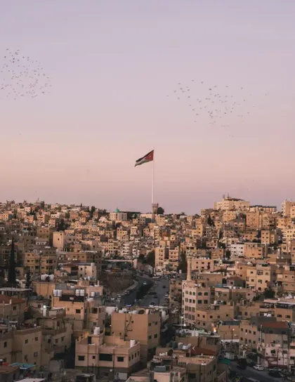 Die Flagge Jordaniens weht über einer dichten Stadtlandschaft aus sandfarbenen Gebäuden unter einem blassrosa Himmel mit fliegenden Vögeln.
