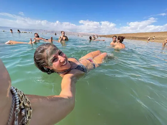 Eine lächelnde Frau mit einer Schlammmaske im Gesicht schwebt im Wasser und macht ein Selfie mit ihrer WeRoad Gruppenreise im Hintergrund.